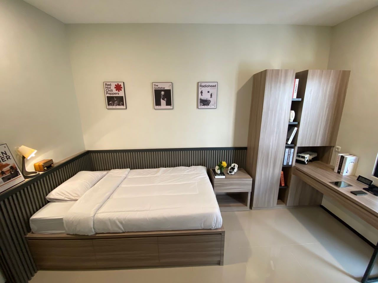 Contoh kamar: kasur putih dengan laci di bawahnya, wainscot bilah kayu abu gelap, lemari dan meja kerja kayu oak, dinding krem sage dengan tiga poster berbingkai