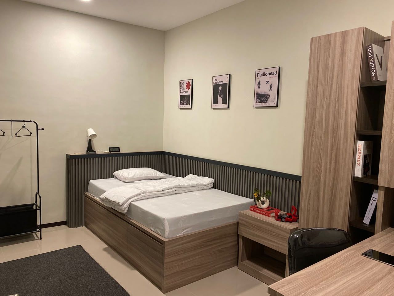 Contoh kamar dari sudut lain: kasur putih di sudut ruangan, rak gantung baju besi hitam, lemari kayu oak tinggi, dan kursi kerja hitam