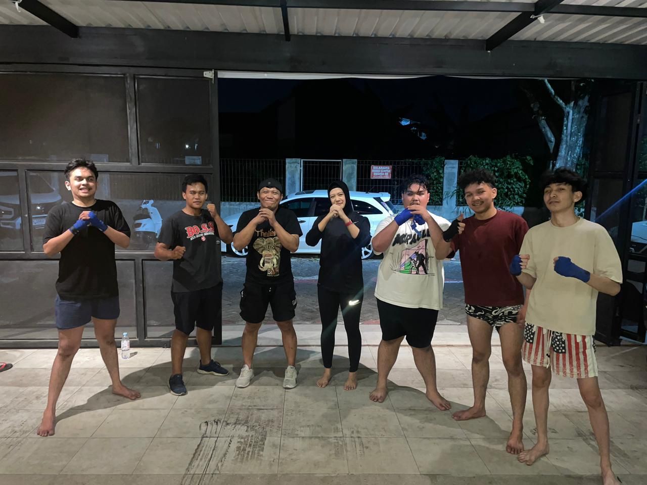 Tujuh penghuni berpose kuda-kuda muay thai dengan sarung tangan di area depan kost pada malam hari
